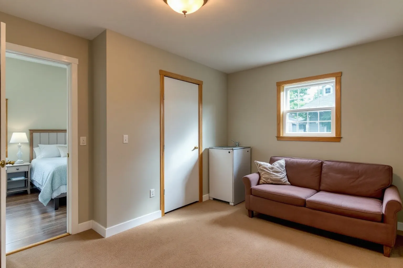 Example of a finished Oshawa basement unit with beige carpet, a mauve sofa under a wood framed window, a bar fridge with a sink and a bedroom through an open door