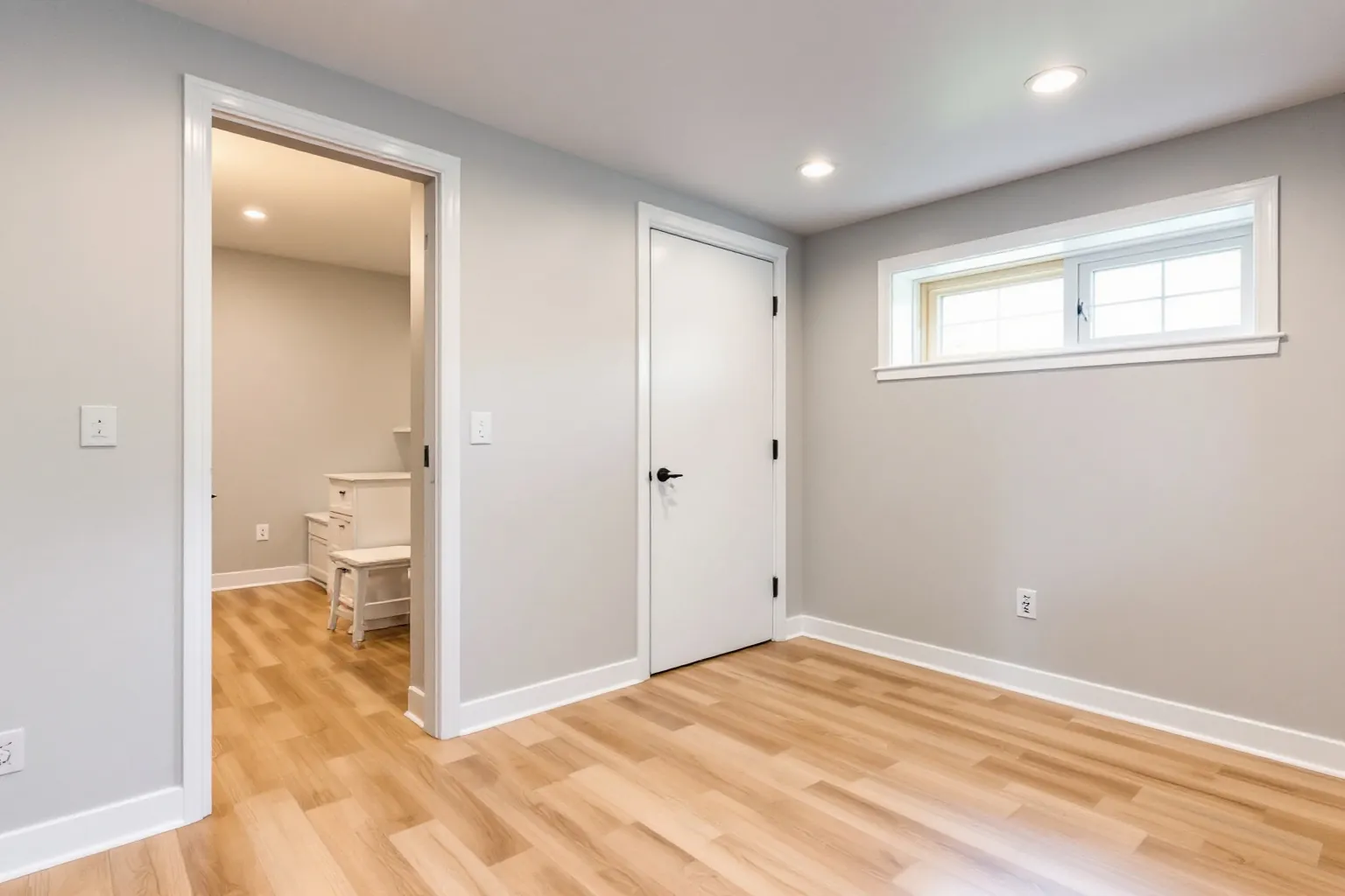 Example of a finished basement bedroom with pale grey walls, light oak plank flooring, a wide window set high on the wall and a doorway to a small office