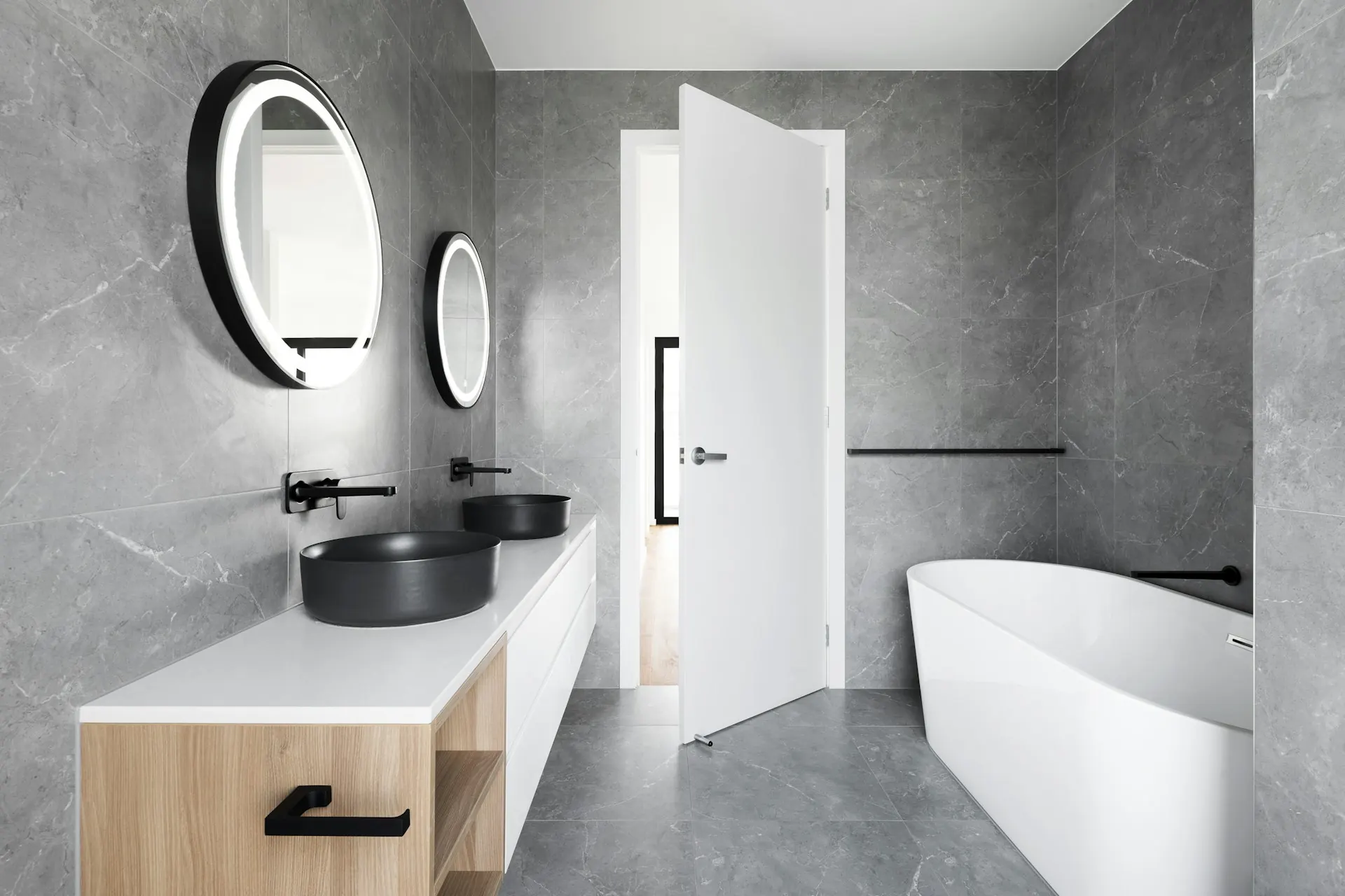 The same bathroom after: grey stone walls, a freestanding tub, a floating vanity and round lit mirrors